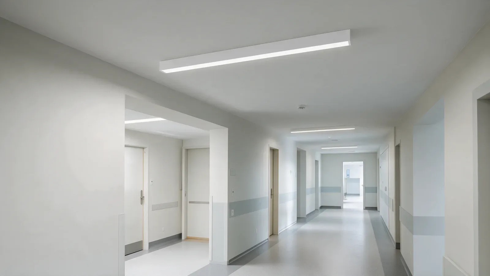 Couloir public d’hôpital avec luminaires linéaires blancs en applique et circulations dégagées.