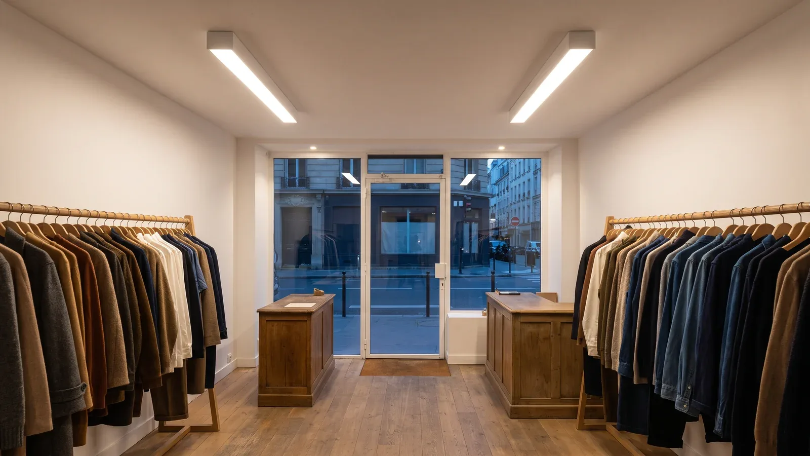 Boutique française avec portants en chêne et lignes lumineuses blanches en applique au plafond, vue vers la vitrine au crépuscule.
